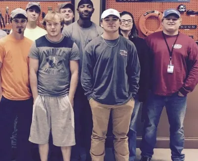 Group of eleven men standing indoors in front of tool displays, casually dressed and smiling at camera
