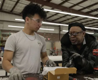 Two workers in a workshop discussing tools at a workbench with power drills and equipment