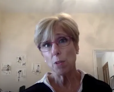 Middle-aged woman with short blonde hair and glasses speaking in a cozy room with wall art and a window.