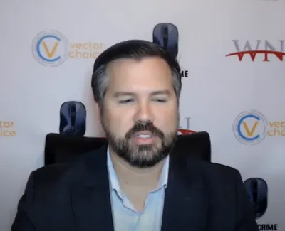 Man with beard and dark blazer speaking in front of a backdrop with Vector Choice and Cyber Crime logos.