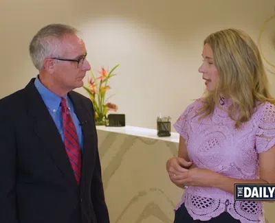 Man in suit and woman in pink lace top converse in modern office lobby with plant and clock.