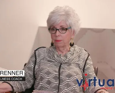 Older woman with white hair and glasses sits in chair wearing zebra-print top, identified as health and wellness coach.