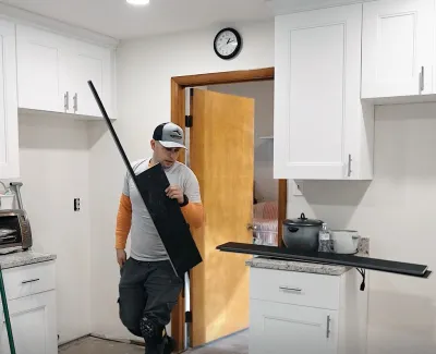 Worker carrying black shelf in kitchen under renovation with white cabinets and wooden door open.