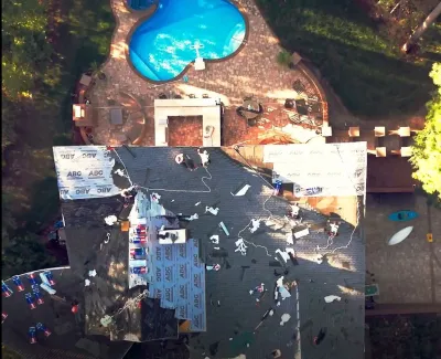 Aerial view of a house undergoing roof repairs next to a uniquely shaped blue swimming pool surrounded by trees.
