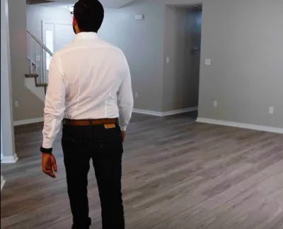 Man walking on light brown vinyl plank flooring inside a modern empty home with gray walls and staircase.