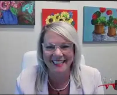 Smiling woman with blonde hair and pearls in front of colorful floral paintings during video call