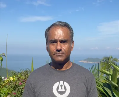 Man in gray shirt with lock logo standing outdoors with ocean, sky, and greenery in the background.