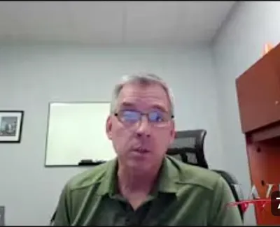 Man in green shirt and glasses speaking during a video call in an office with a whiteboard and wooden cabinet.