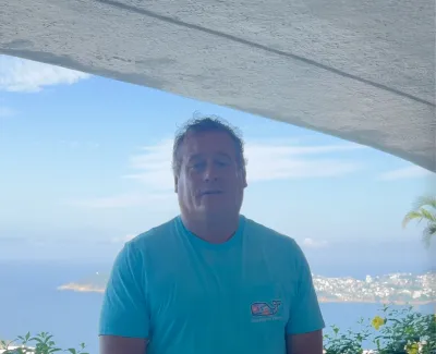 Man in blue t-shirt stands under a white roof with ocean and hillside cityscape in the background