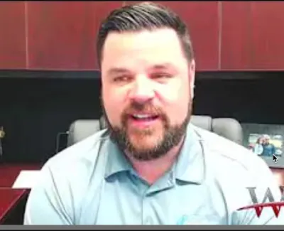 Man with beard wearing a light blue shirt speaking in a home office with wooden cabinets behind him