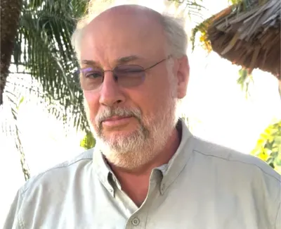 Bearded man wearing glasses and a light-colored button-up shirt outdoors near greenery and thatched roof.