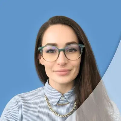 Portrait of a smiling woman wearing glasses, striped shirt, and gold necklace against a blue background.