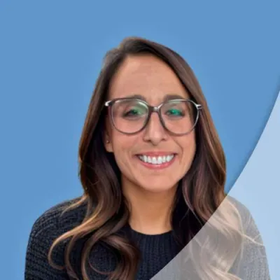 Smiling woman with long brown hair and glasses wearing a black sweater against a blue background.