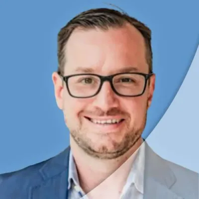 Smiling man with glasses wearing a blue blazer and checkered shirt against a blue background.