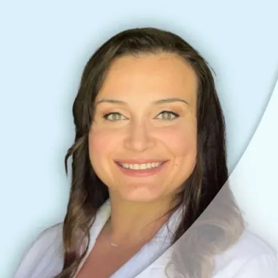 Smiling woman with long dark hair wearing a white coat against a light blue background.