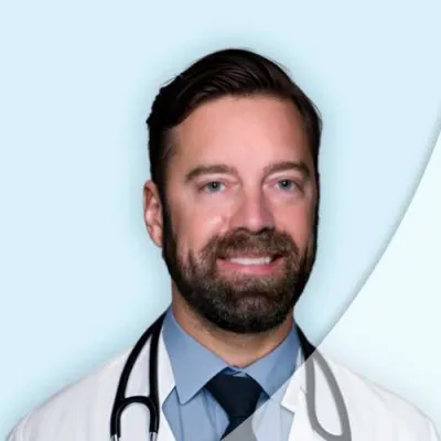 Smiling male doctor with beard wearing white coat and stethoscope on light blue background