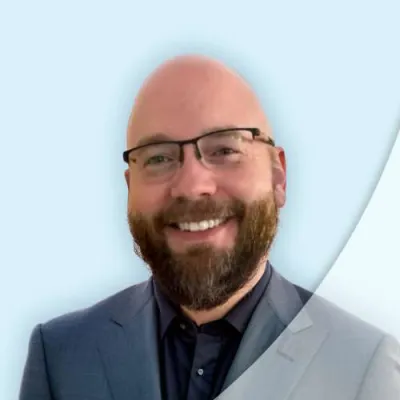 Smiling bald man with glasses and beard wearing a gray suit and black shirt against light blue background.
