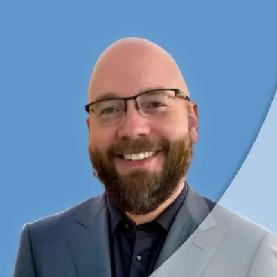 Smiling bald man with glasses and beard wearing a blue suit and dark shirt against a blue background