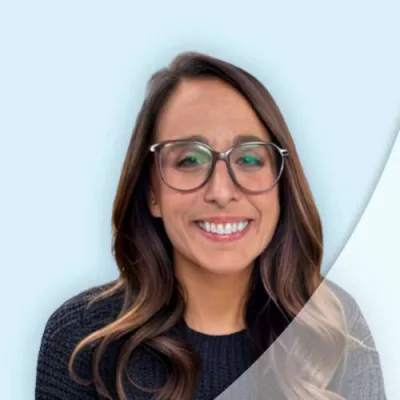 Smiling woman with glasses and long wavy hair wearing a black sweater against a light blue background.