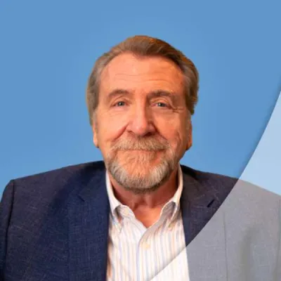Smiling older man with gray hair and beard wearing a blue blazer and striped shirt against a blue background.
