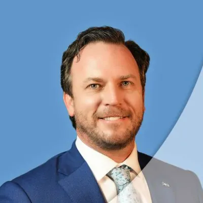 Professional man in blue suit and patterned tie smiling against a blue background with geometric shapes.