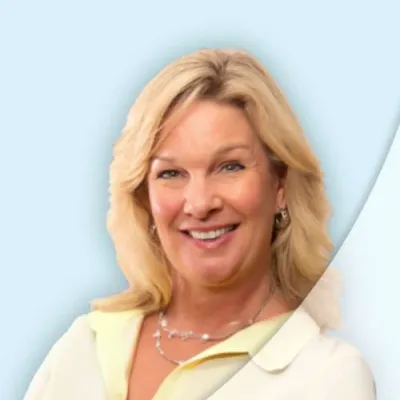 Smiling blonde woman in light yellow blouse with silver necklace and earrings on pale blue background.