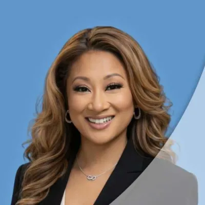 Smiling woman with long wavy hair wearing a black blazer and silver necklace against a blue background