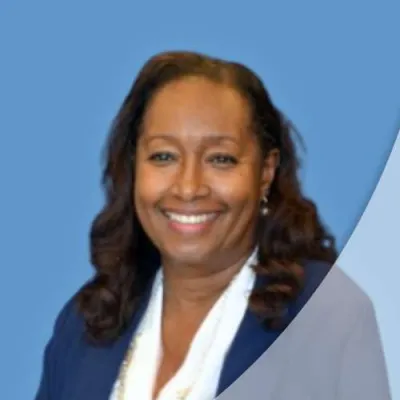 Smiling woman with dark hair wearing pearl necklace and navy blazer against light blue background