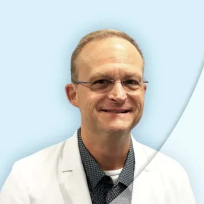 Smiling middle-aged man wearing glasses, a white coat, and a patterned shirt on a light blue background
