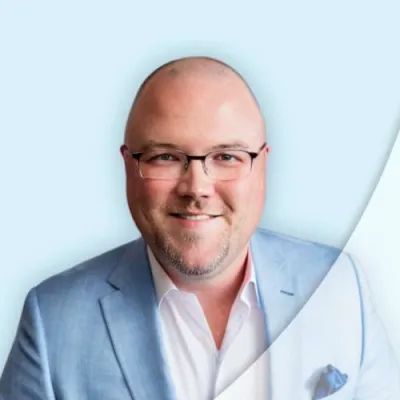 Smiling bald man with glasses wearing a light blue suit jacket and white shirt against a light background.
