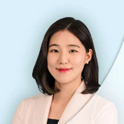 Professional female healthcare provider in white coat with dark hair smiling against light blue background.