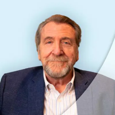 Smiling older man with beard in navy blazer and striped shirt against light blue background