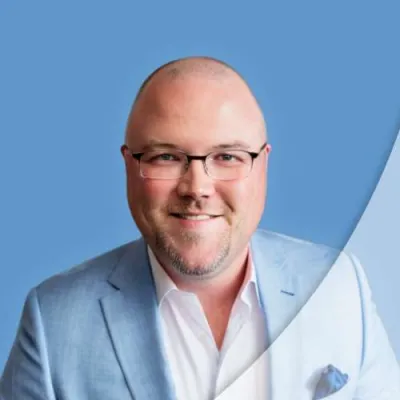 Smiling bald man wearing glasses and a light blue suit jacket against a blue background