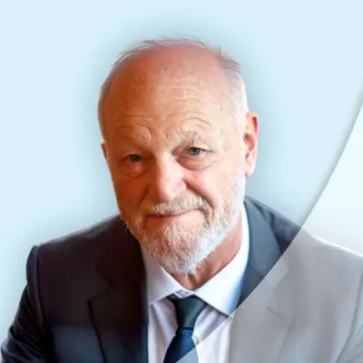 Portrait of an elderly man in a suit with a white beard and blue tie against a light blue background.