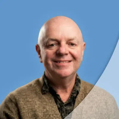 Smiling bald man wearing a brown sweater and patterned shirt against a blue background with curved overlay.