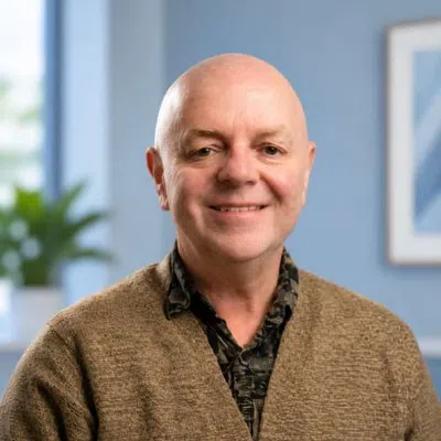 Smiling bald man wearing a brown sweater and patterned shirt in a softly lit indoor background.