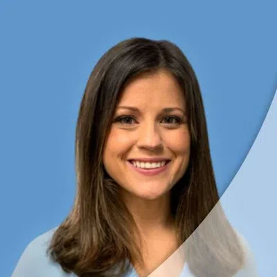 Smiling woman with shoulder-length brown hair wearing a light blue blouse against a blue background.