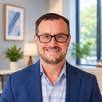 Smiling man with glasses wearing a blue blazer and checked shirt in a bright modern office setting.