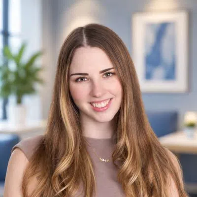 Young woman with long wavy hair smiling in a softly focused indoor setting with plants and blue decor.