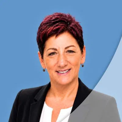 Professional woman with short red hair and black blazer smiles against a blue and light grey background.