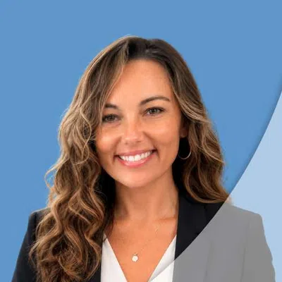 Smiling woman with wavy brown hair wearing a black blazer and white top against a blue background.