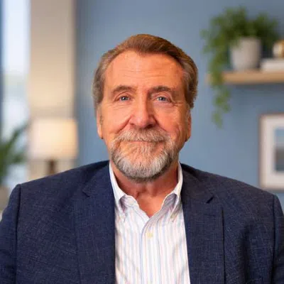 Smiling older man with gray beard wearing a navy blazer and striped shirt in a cozy room with plants and lamp.