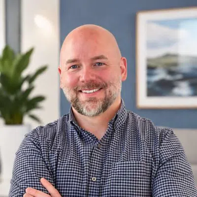 Smiling bald man with beard in checkered shirt poses with crossed arms indoors with blurred plant and painting background.