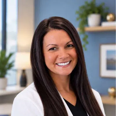 Smiling woman with long dark hair wearing a white blazer and black top in a modern office setting.