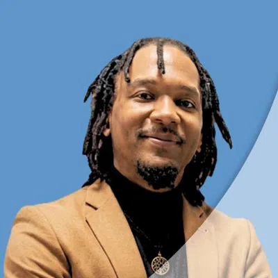 Man with dreadlocks wearing tan blazer and black turtleneck against a blue background smiling confidently.