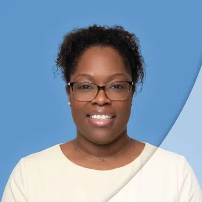 Smiling woman with glasses and curly hair wearing a white top against a blue background.