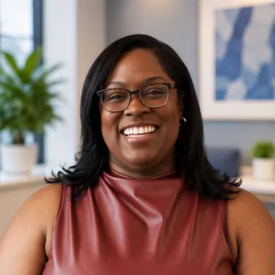 Smiling woman wearing glasses and a sleeveless maroon top in a well-lit modern office with plants and artwork.