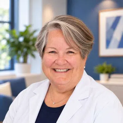Smiling older woman with short gray hair wearing a white coat in a bright office with blue walls and plants.