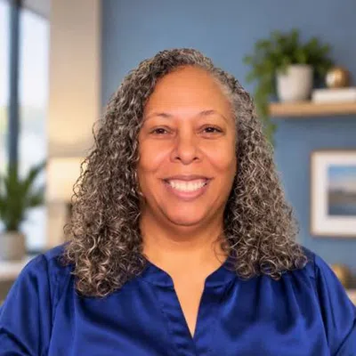 Smiling woman with curly gray hair wearing a blue blouse in a modern office setting with blurred background.