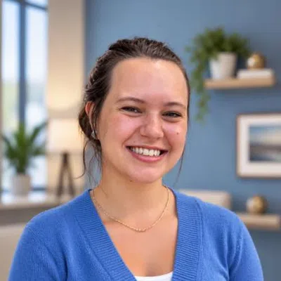 Smiling woman with dark hair in blue cardigan standing in a warmly lit modern office space.